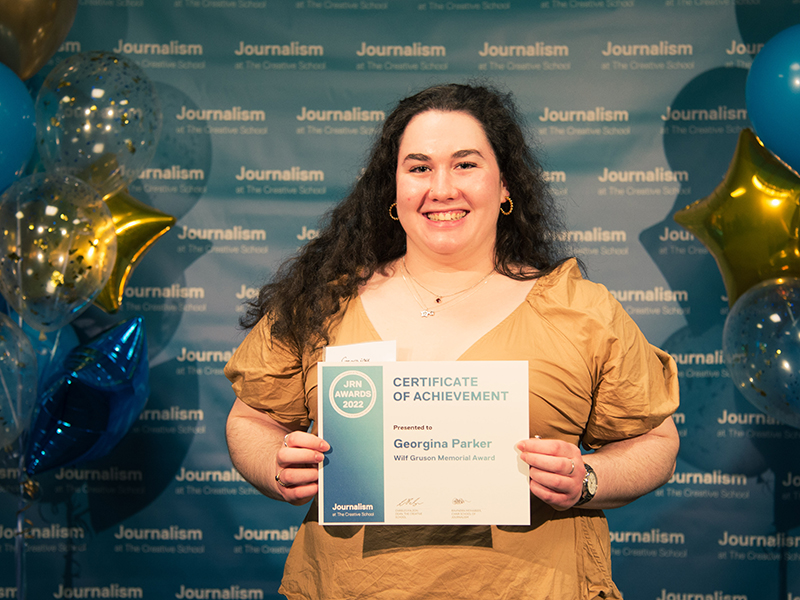 Georgina Parker holds a certificate while standing in front of a blue backdrop that repeats, "Journalism at The Creative School."