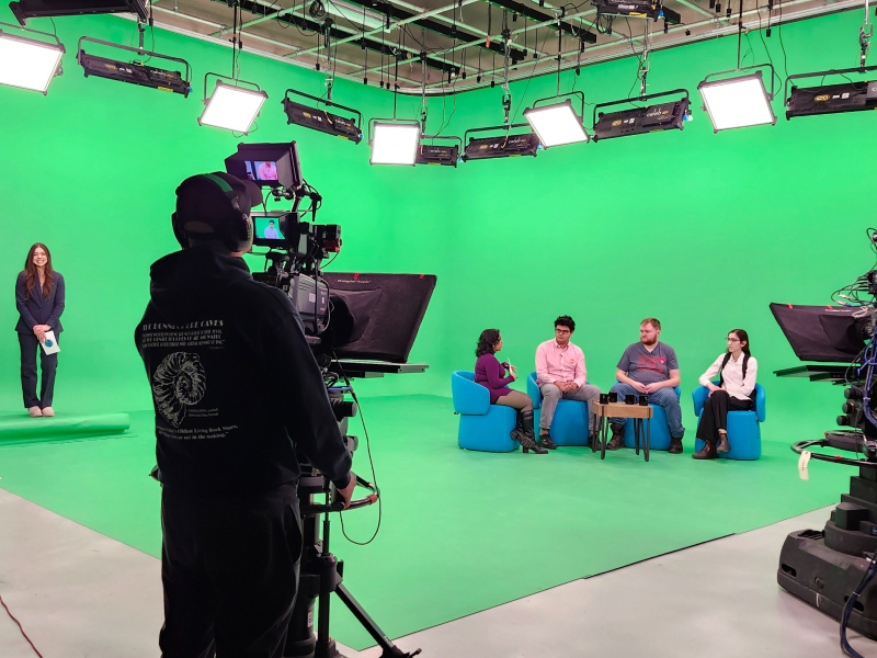 In a green screen studio four people sit together in conversation while being filmed on camera