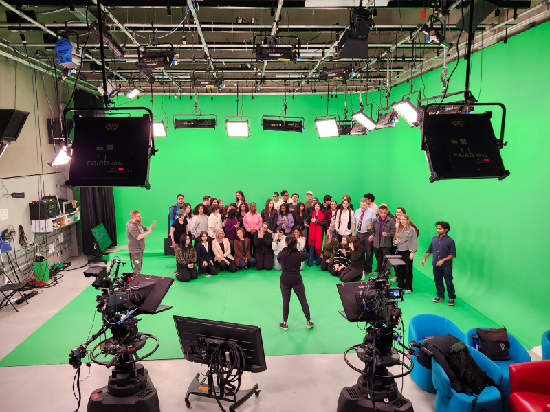 A crowd of people stand together in front of a green background in a studio.