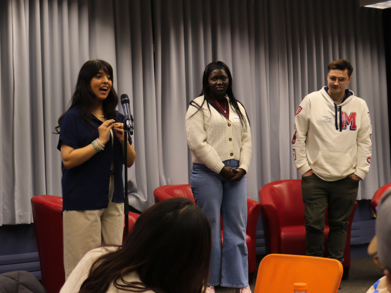 Three people stand next together on a raised platform while one speaks into a microphone.