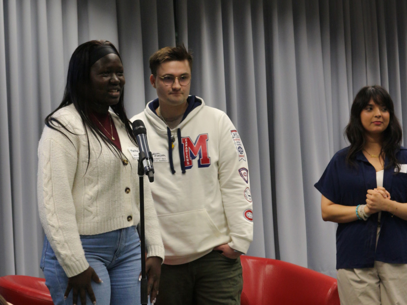 Three people stand next together on a raised platform while one speaks into a microphone.