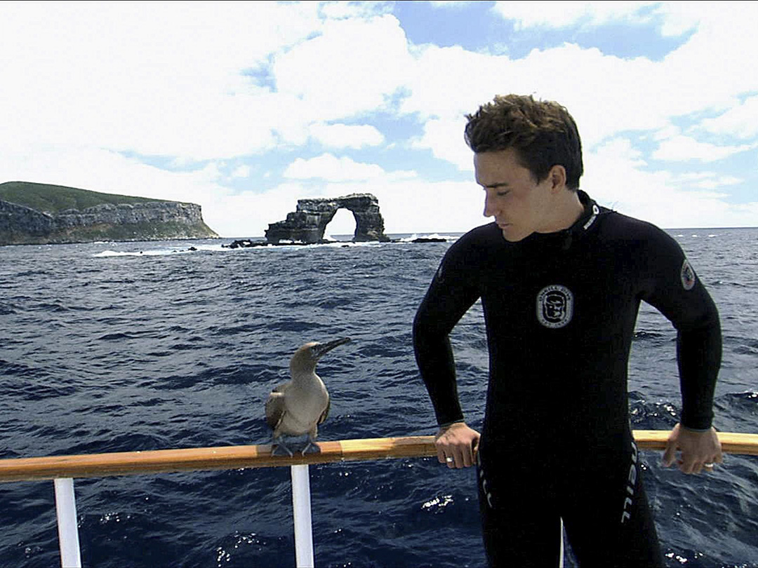 Still of Rob Stewart from Sharkwater (2006). He is on a boat in the Galapagos looking at a blue footed boobie.