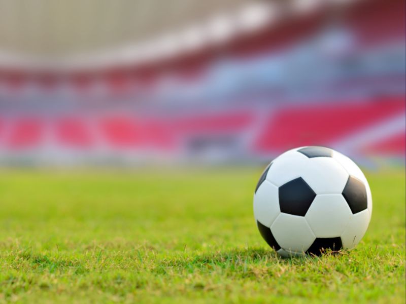 A soccer ball on the grass with a stadium in soft focus in the background. 