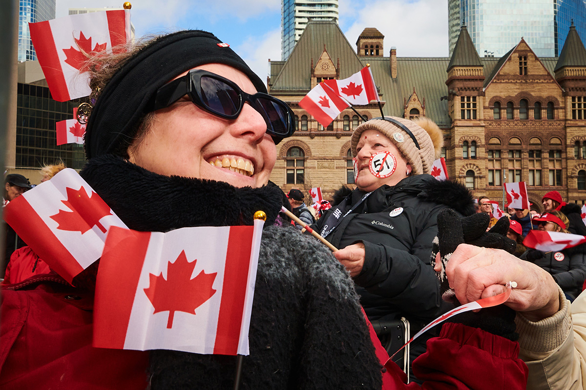 A close up shot of a person smiling while waving a Canadian flag with more flags and people in the background.