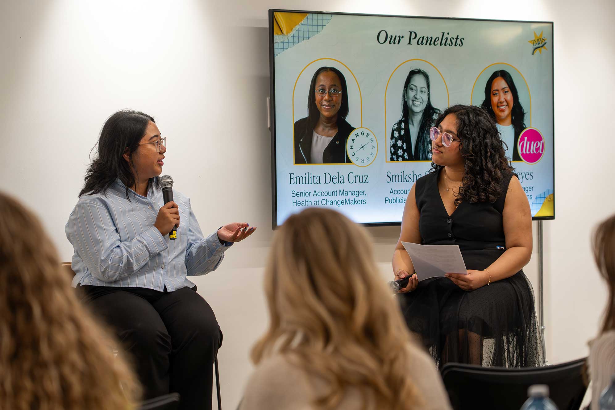 Two people sit on high chairs in front of crowd while speaking on a panel