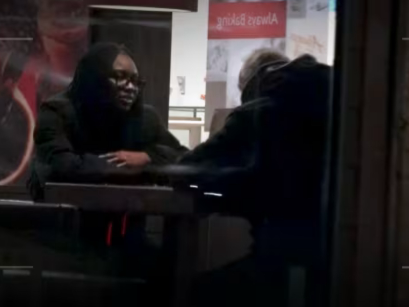 A photograph taken outside of a Tim Hortons looks through the glass to see two people sitting across from each other having a conversation.