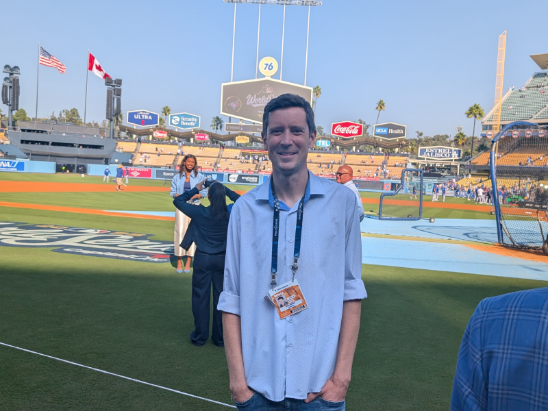 Male alumni stands on Dodgers Stadium field while smiling at the camera.