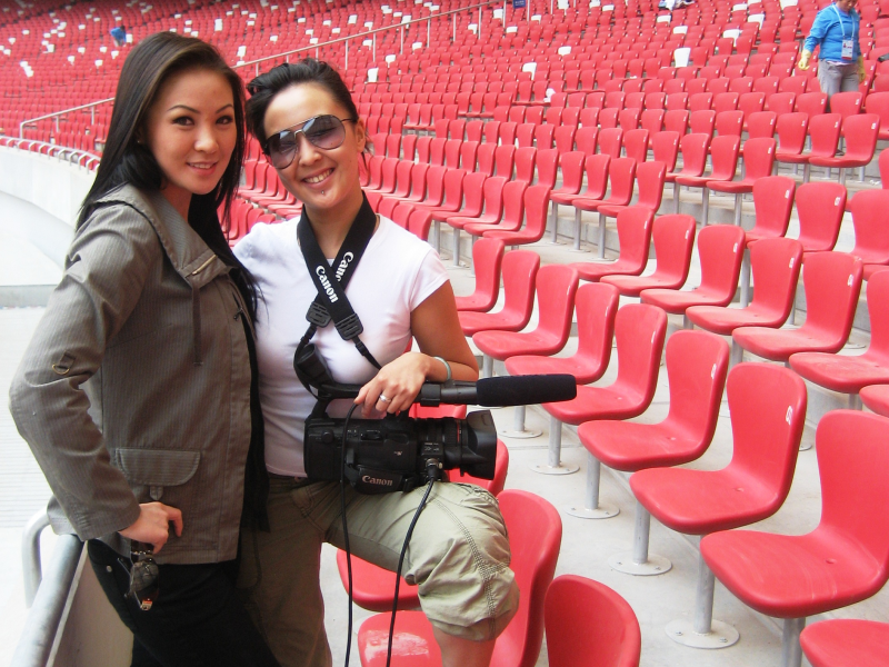 Two people stand in rows of chairs with a camera smiling at the camera.