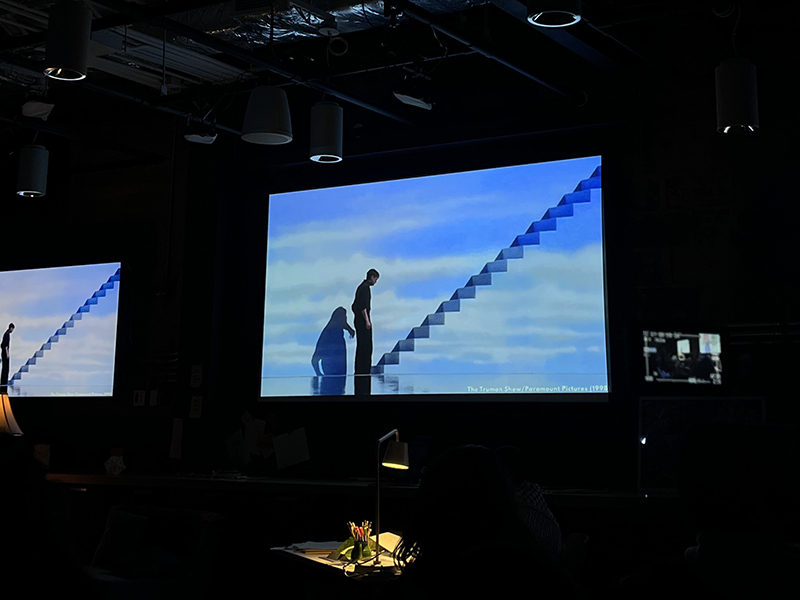 A person stands in front of a blue sky projection with stairs leading upwards.