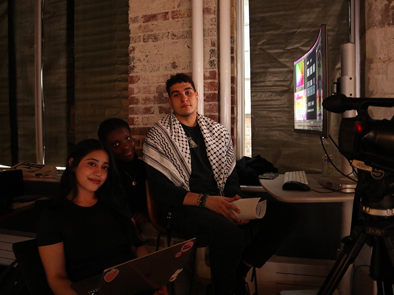 Three students smile at the camera while sitting down looking at a computer.