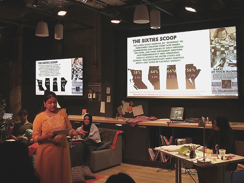 A student stands up while reading a card during a performance with people sitting behind her and a projection on the wall.