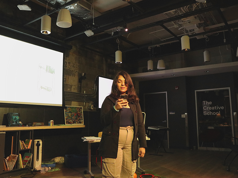 A student stands up while reading off her phone during a performance with a projection on the wall behind her.