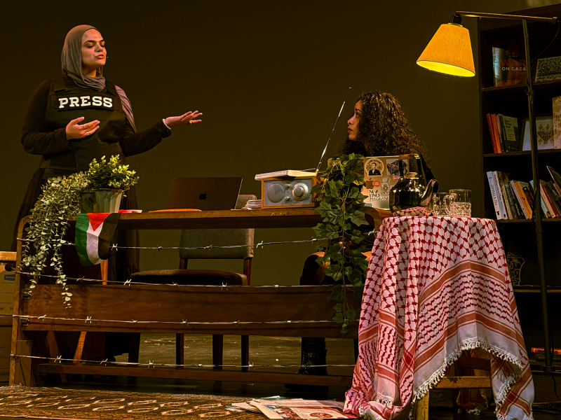 A photo of an on stage performance with two performers talking to each other behind a table