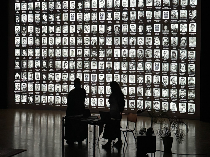 Two people sit in the dark while a background with peoples faces fill the wall behind them