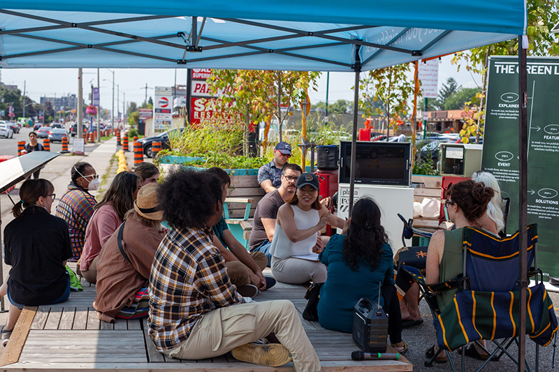 A group of people sit under a blue shade tent facing Anita Li as she gives a speech at an event.