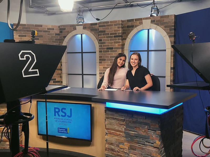Two students sit behind a news desk smiling at the camera.