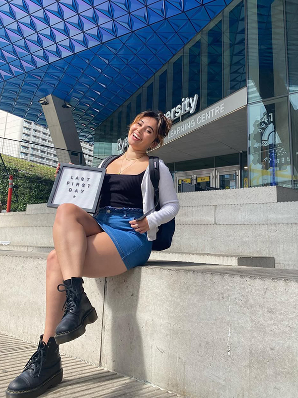A student holds a sign that says, "Last first day," while siting on concrete steps in front of a blue building.