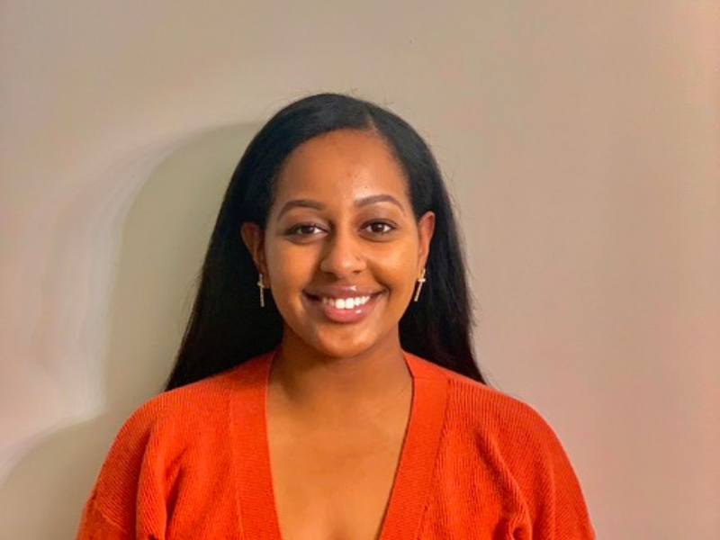 Head shot of journalism student Bana Yirgalem standing against a beige background and wearing an orange shirt. 