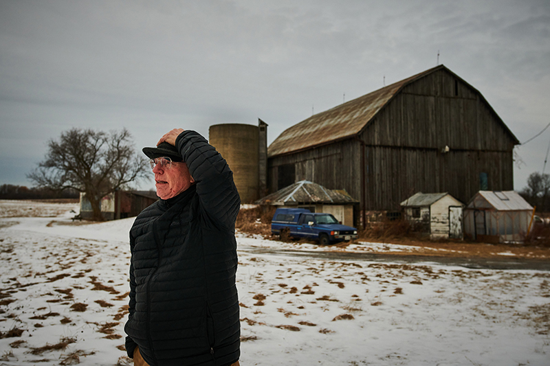 A photo of a person looking to the side while standing in front of a barn in winter.