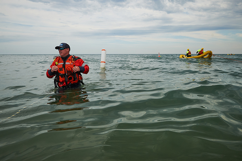 A person in a red life jacket stands in the water with a boat in the background.