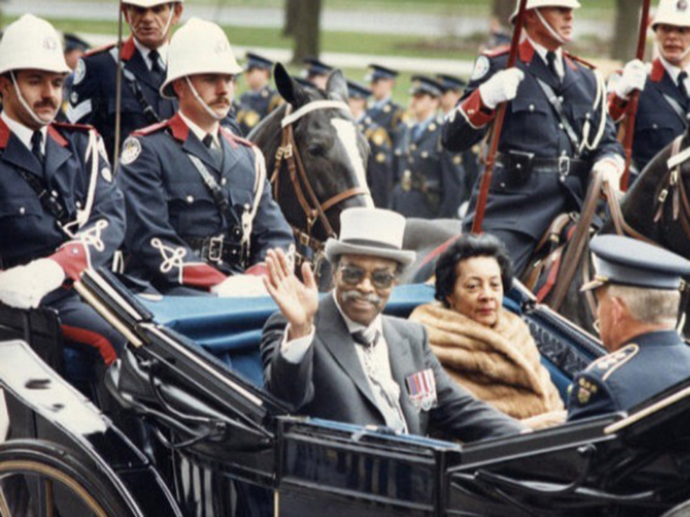 The Honourable Lincoln Alexander greeting the crowd with his wife on a car, surrounded by the RCMP