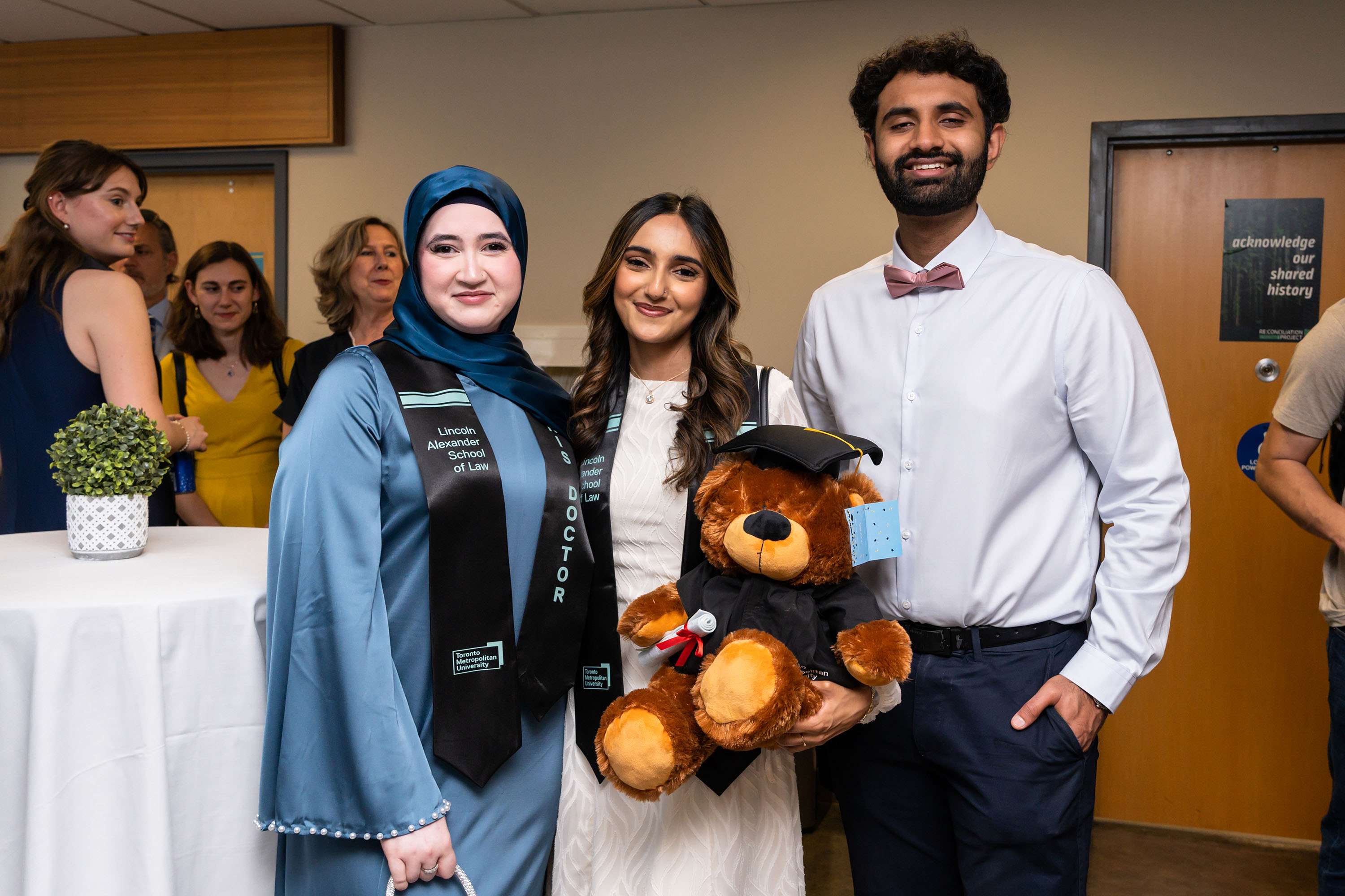 grads pose for photo with a stuffed bear wearing a graduation cap and gown