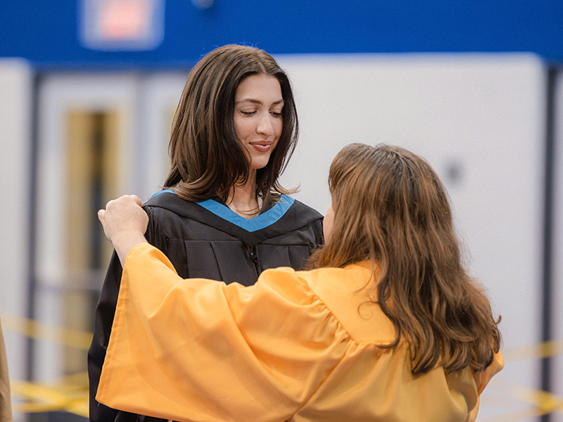 A woman in a yellow robe adjusts a graduating students regalia