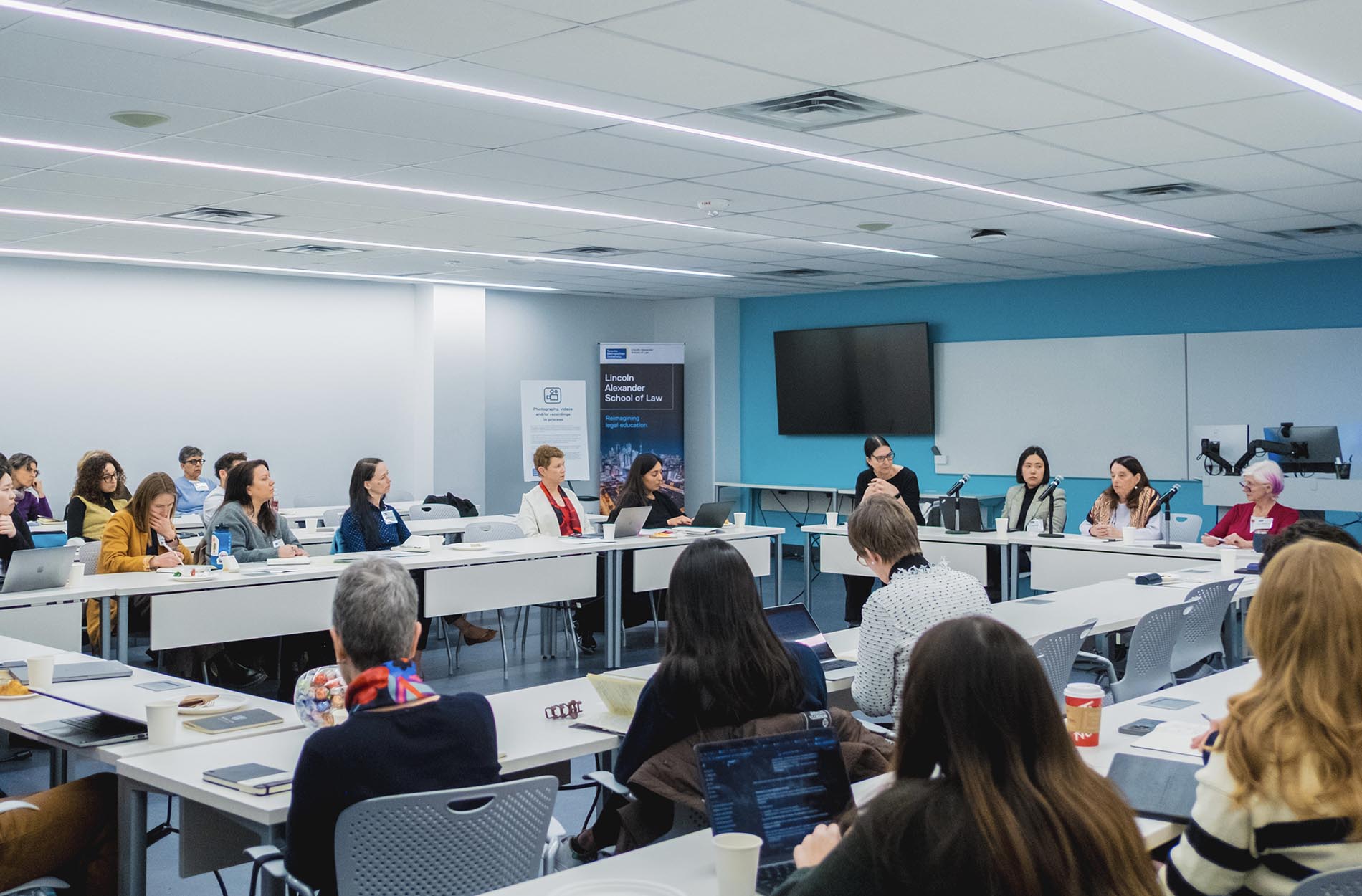 Attendees sit at U-shaped tables participating in a conference workshop inside a bright, modern classroom