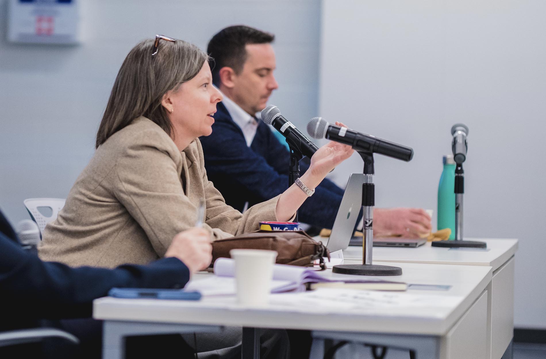 A woman gestures and speaks into a microphone while seated at a panel table during a conference