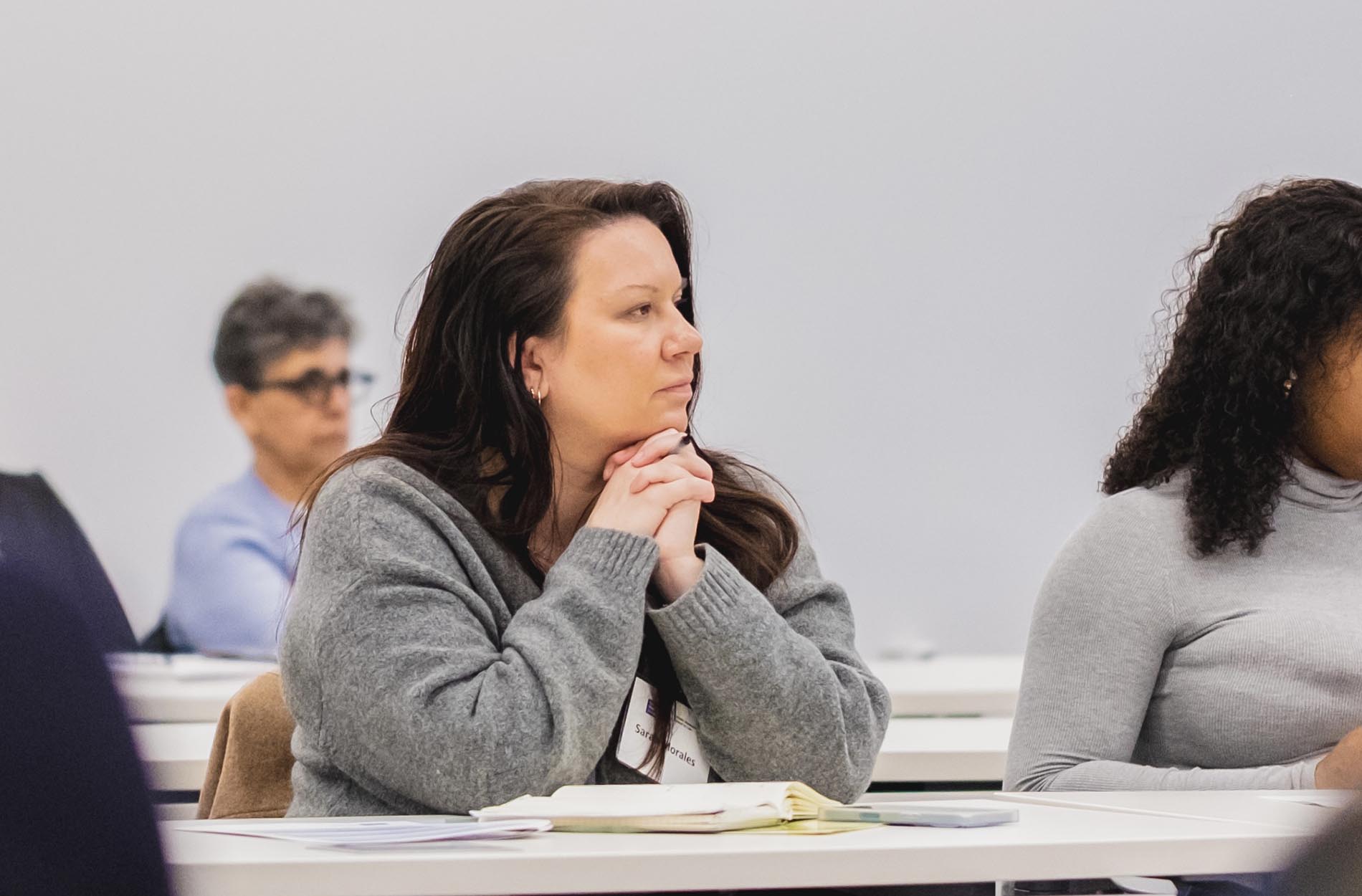 A woman in a grey sweater sits with her hands clasped under her chin, listening attentively during a conference session