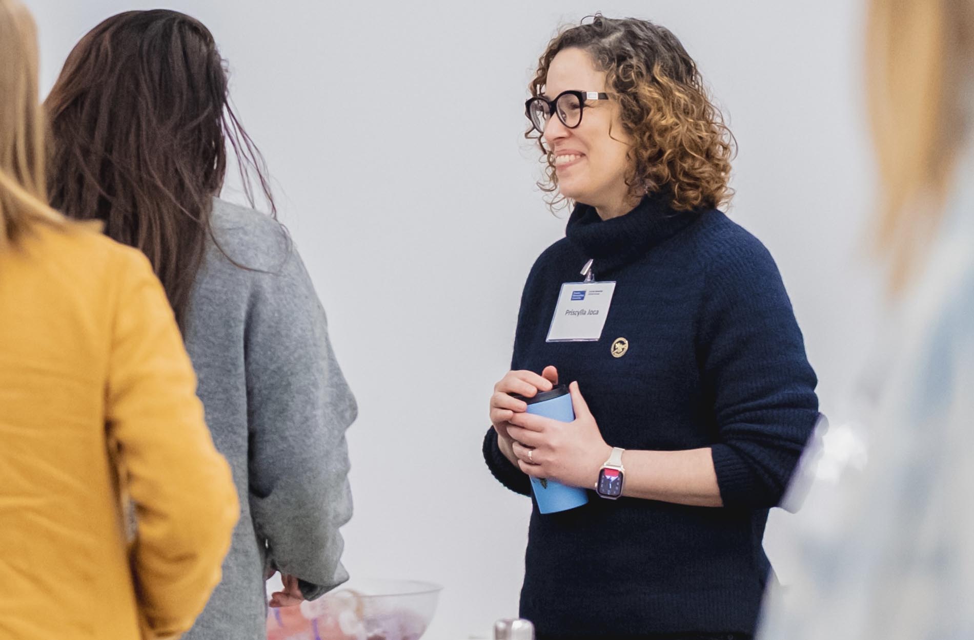 A woman wearing glasses and a navy sweater smiles while holding a blue travel mug and conversing with other attendees