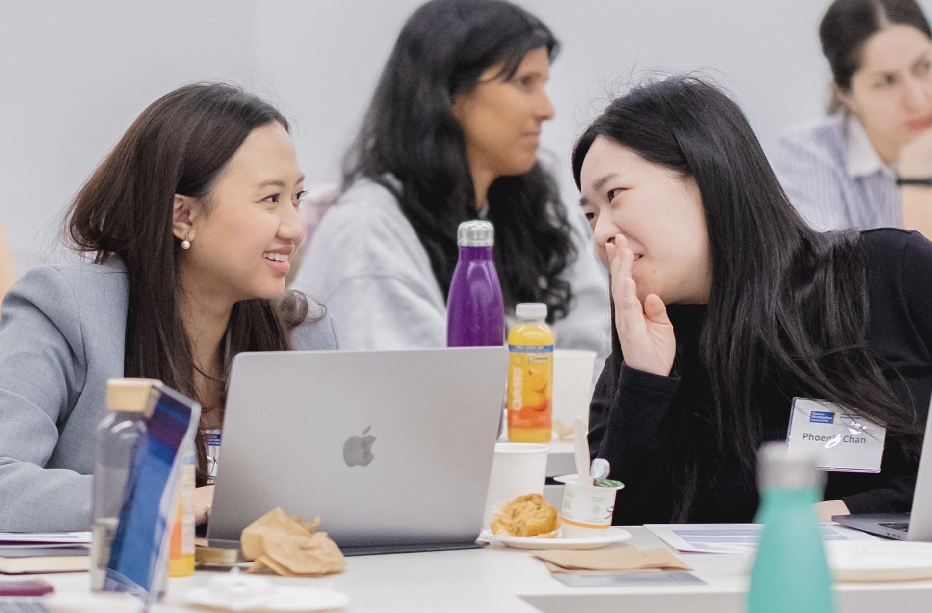 Two women are smiling and talking together at a table with laptops and beverages during the conference workshop