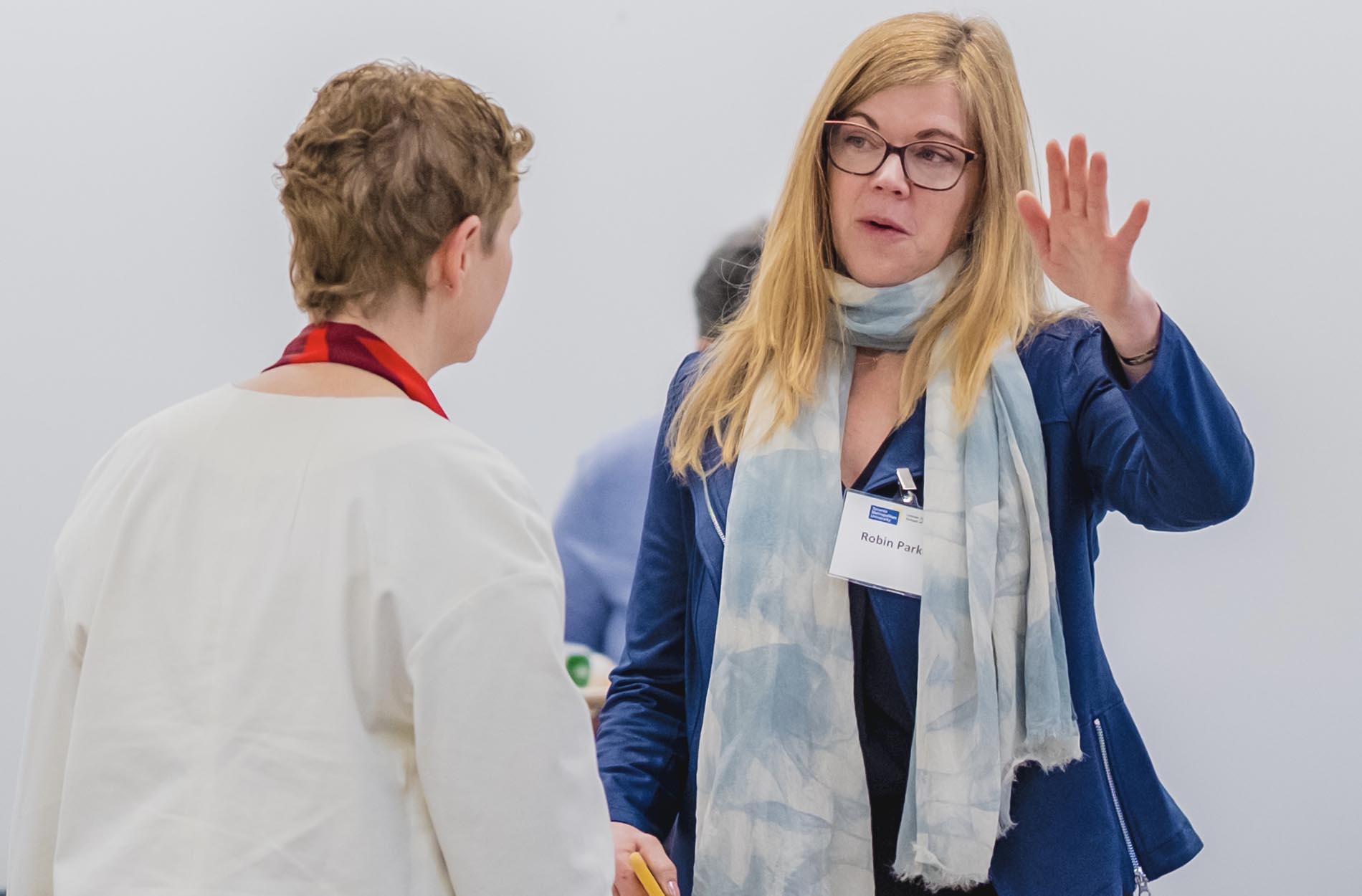 A blonde woman wearing a blue jacket and a scarf raises her hand while speaking to another attendee at a conference