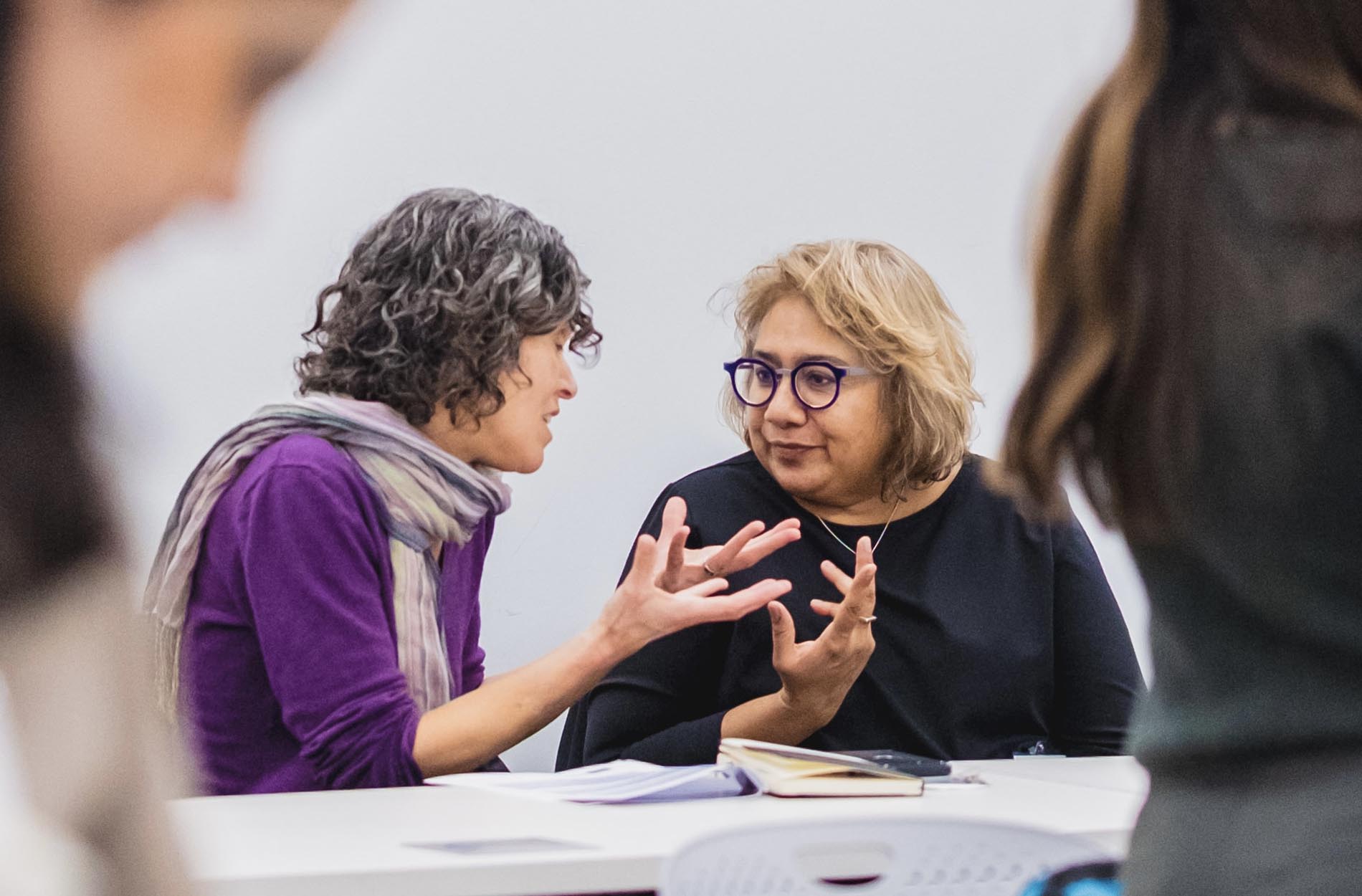 A woman in a purple shirt gestures expressively with her hands while speaking with another woman wearing purple glasses at a table