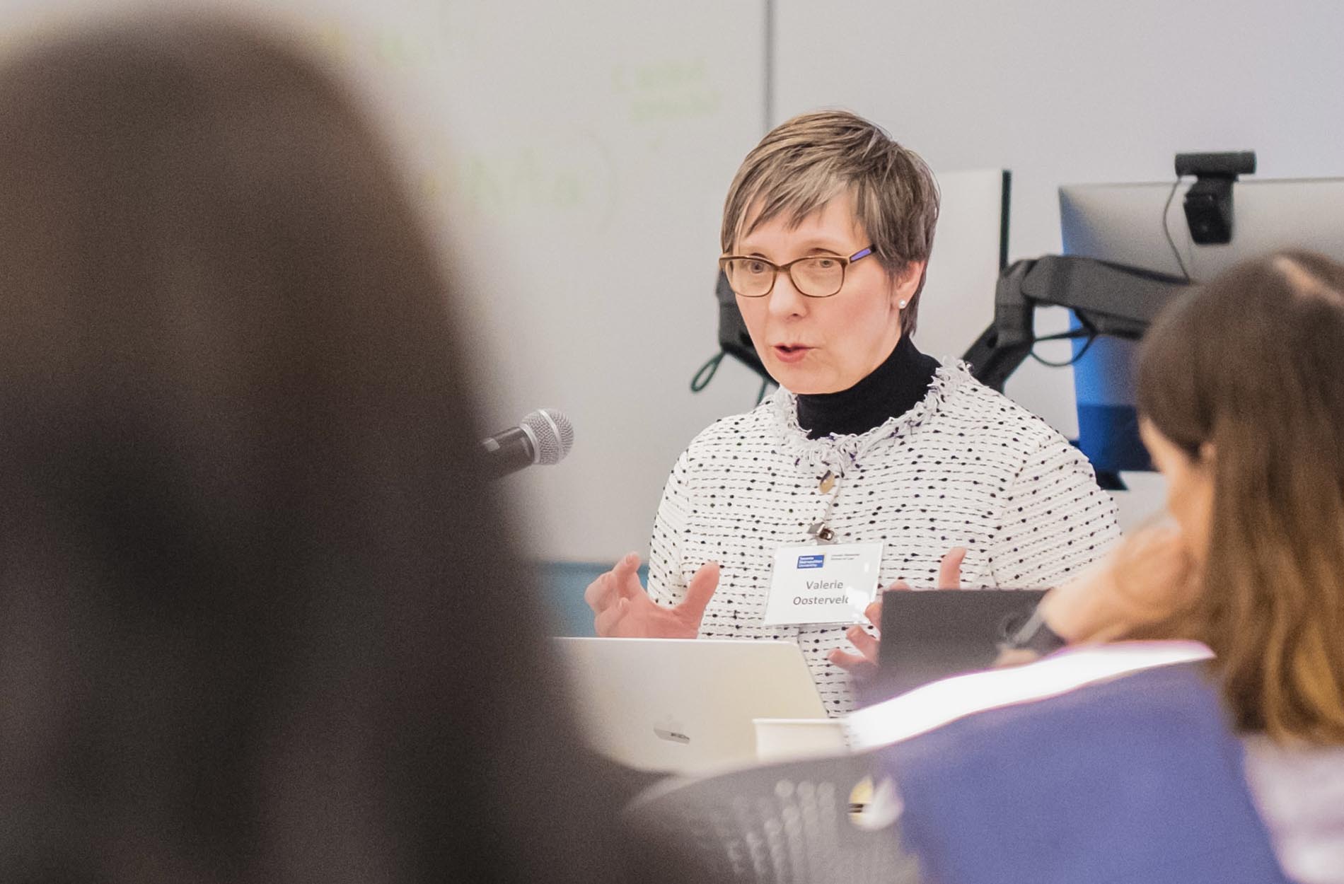 A woman with short hair and glasses wearing a patterned sweater gestures with her hands while speaking at a conference presentation