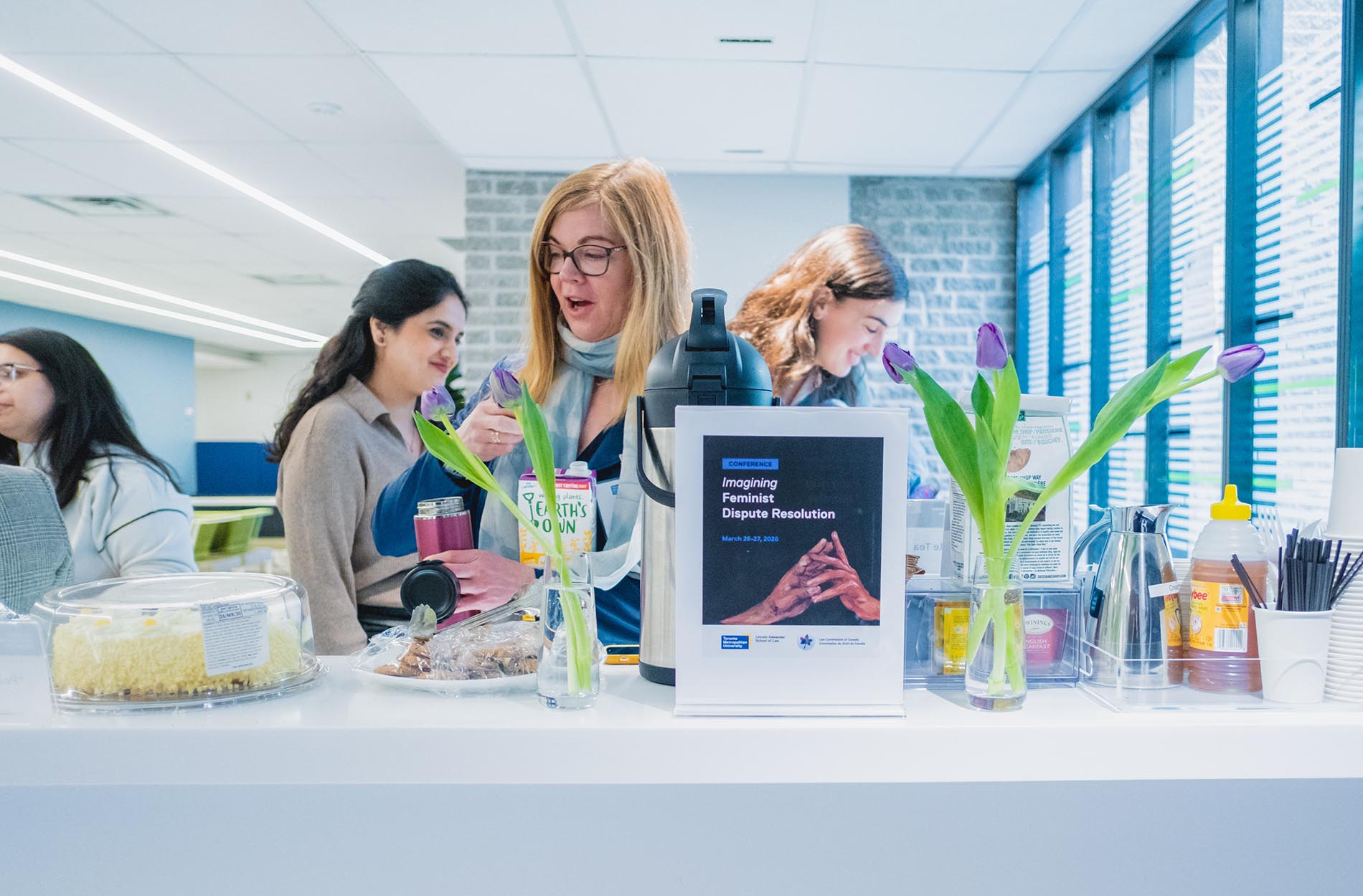 Attendees gather around a refreshments counter featuring coffee, cake, purple tulips, and an "Imagining Feminist Dispute Resolution" sign