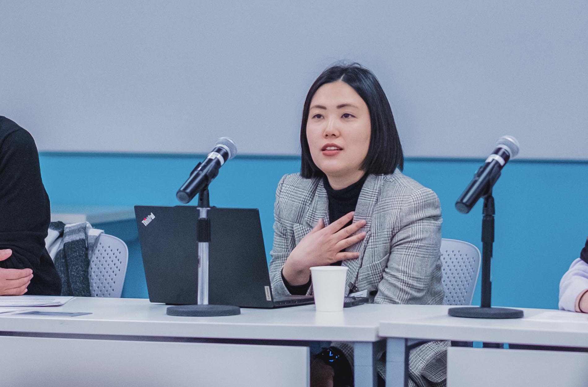 A woman in a grey plaid blazer speaks with her hand on her chest while seated at a panel table with a laptop and microphone