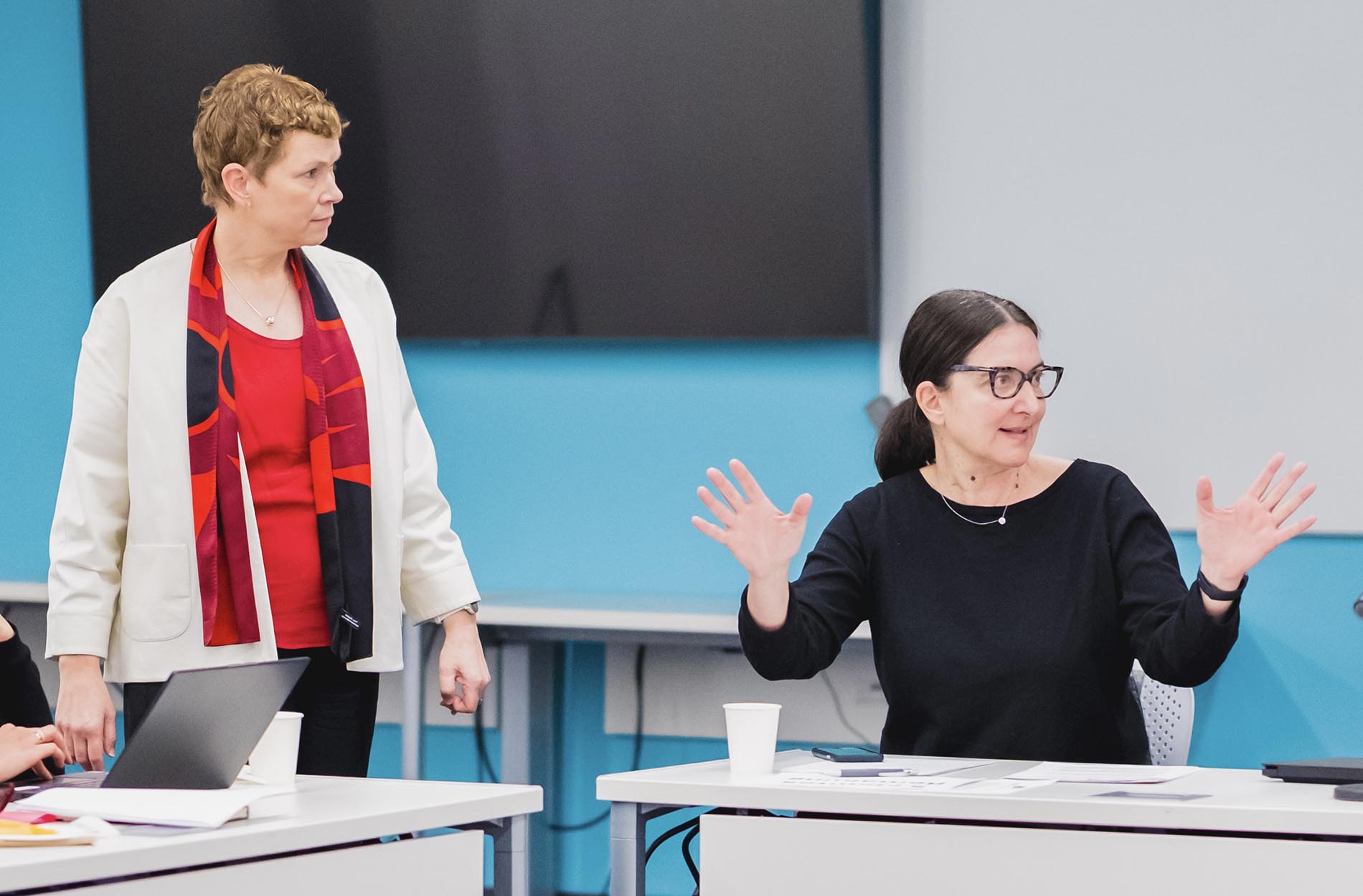A seated woman in a black top gestures expressively with both hands while speaking next to a standing colleague in a white blazer and red shirt