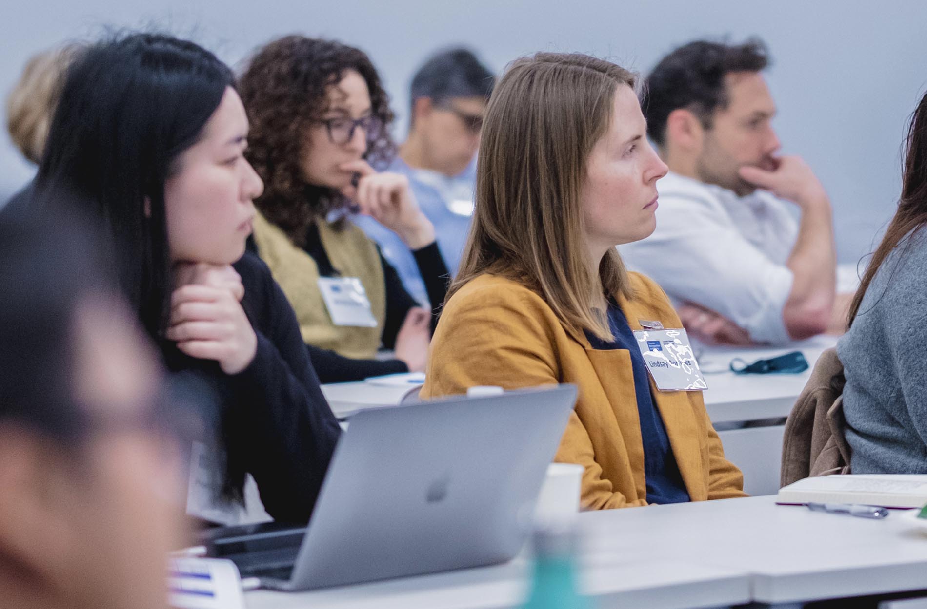 A woman in a mustard yellow blazer sits at a desk with a laptop, listening attentively alongside other attendees during a presentation