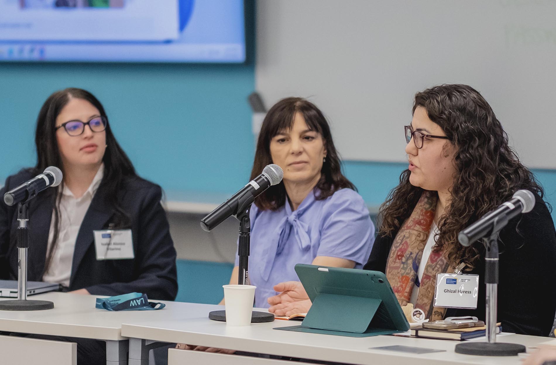 Three women sit at a panel table during a conference, with the woman on the right speaking into a microphone beside her tablet