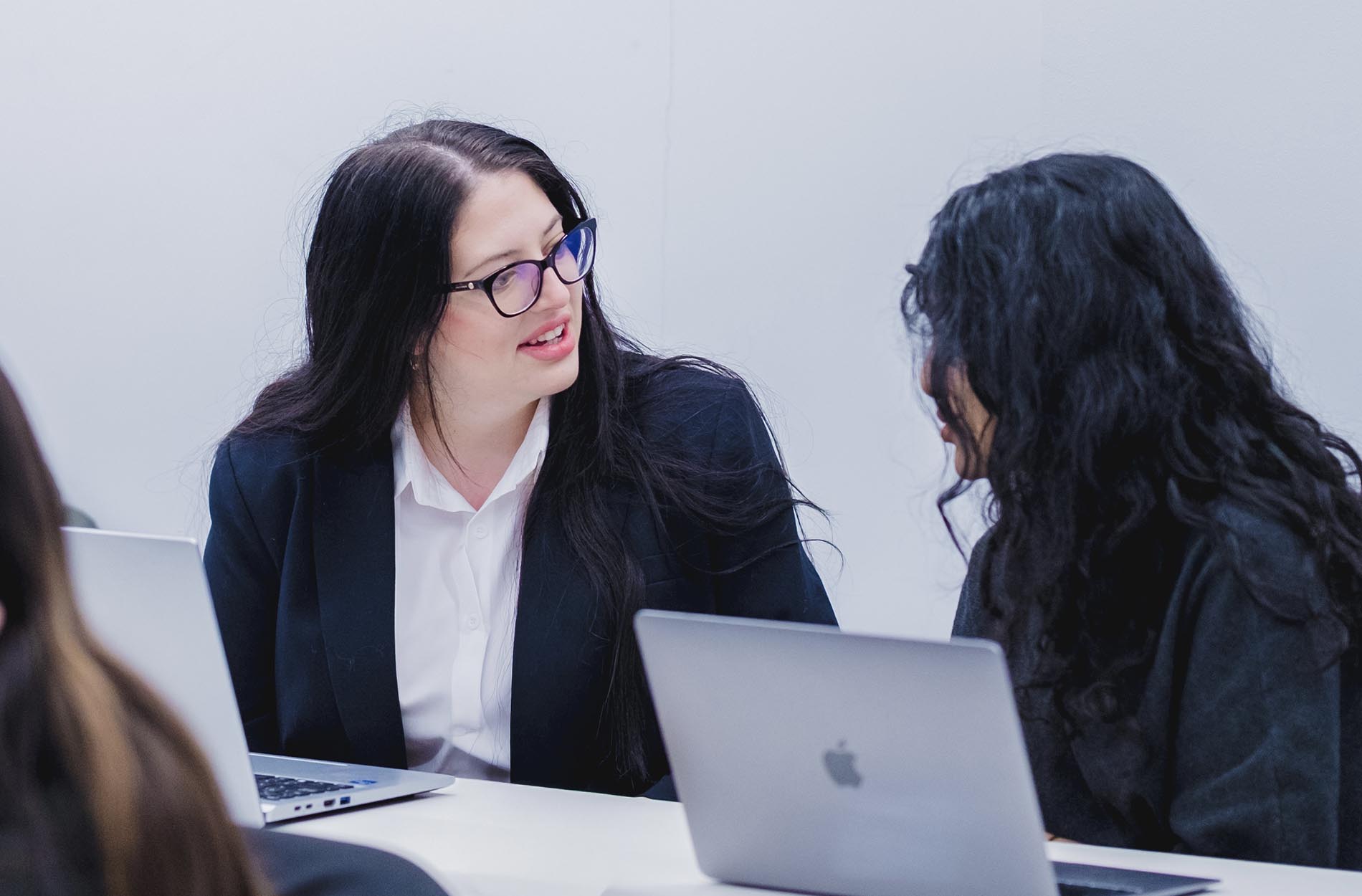 A woman in a black blazer and glasses smiles while conversing with a colleague at a table with laptops