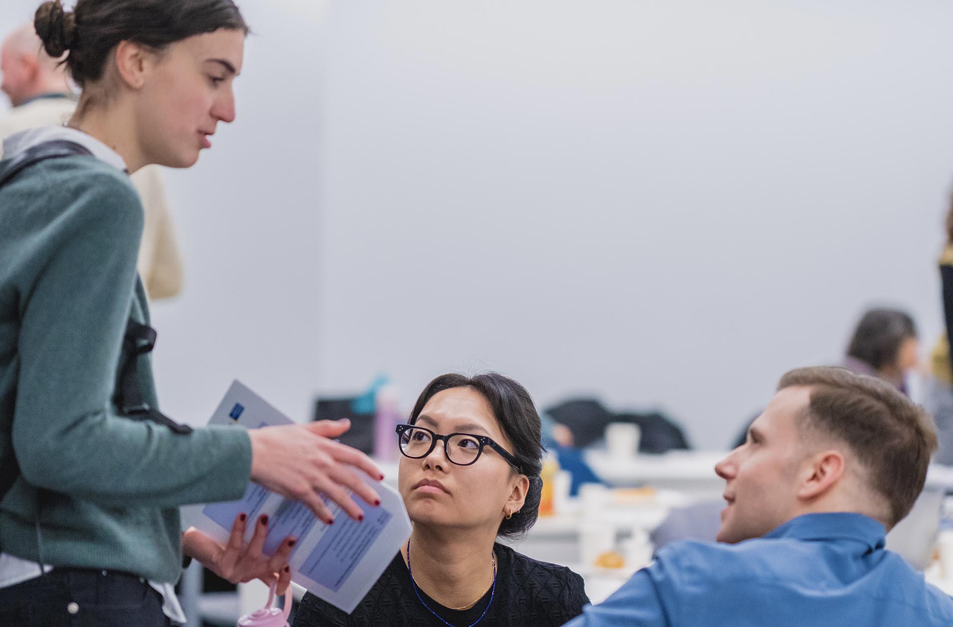 A standing woman in a green sweater holding a document speaks to two seated attendees who are looking up at her