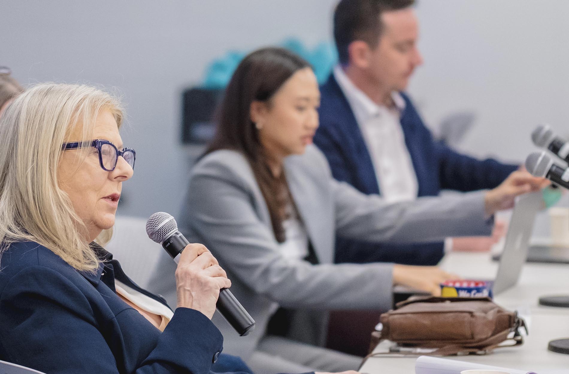 A blonde woman wearing glasses speaks into a microphone while seated at a panel table alongside other presenters working on laptops
