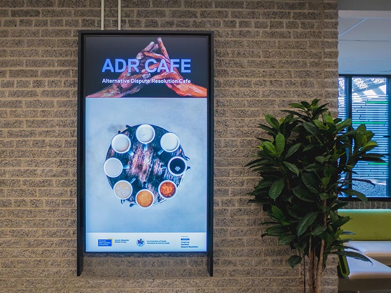 A digital screen mounted on a brick wall displays a poster for the "ADR CAFE" featuring an overhead image of coffee mugs arranged in a circle