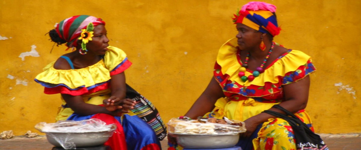Two women street vendors wearing colourful dresses and talking to each other.