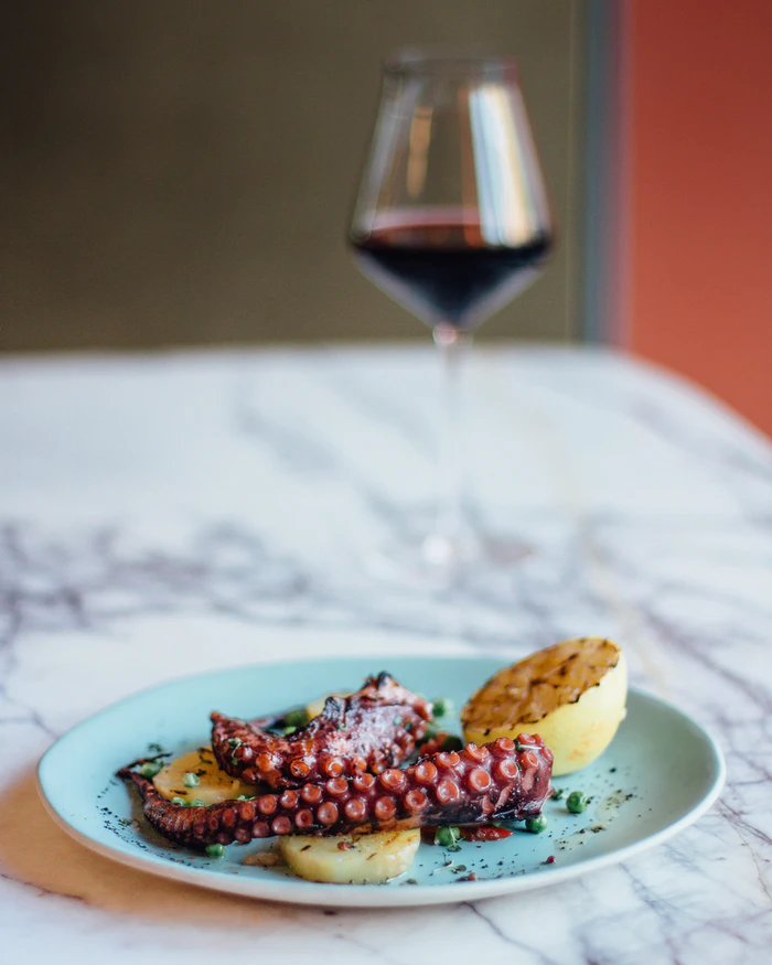 A dish consisting of grilled octopus and lemon, and a glass of wine, sitting on a dining table.