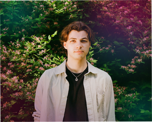 "A portrait of Aidan Wolfson looking forward against a dense backdrop of green bushes and blooming pink flowers. He is wearing a black t-shirt with a silver necklace under an unbuttoned light grey shirt. The image features a purple colour gradient overlay on the upper right side and a soft green hue on the bottom left." 