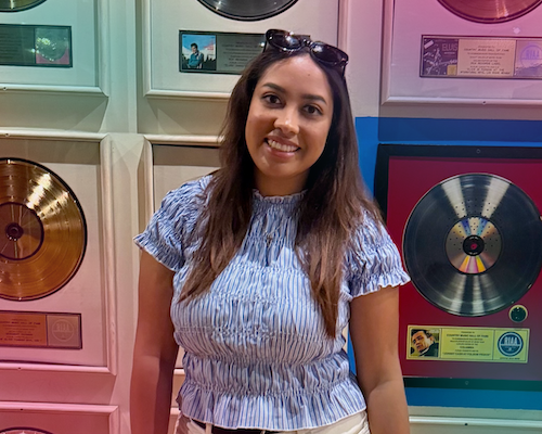 "A portrait of Jaylin Evans smiling with sunglasses resting on her head, wearing a light blue and white striped short-sleeved top. She is standing in front of a wall lined with framed gold and vinyl music records."