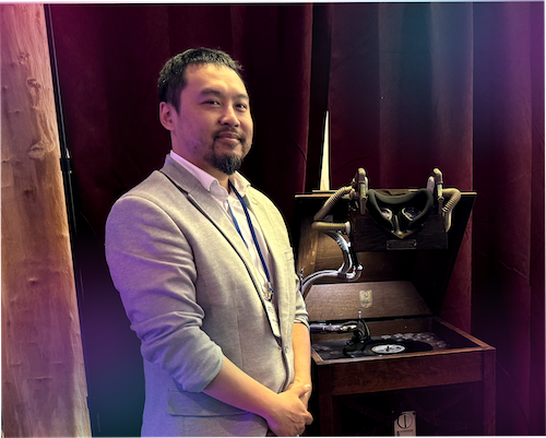 "A portrait of Jeffrey Tran looking toward the camera, wearing a light grey blazer over a white shirt with an event lanyard. He is standing next to a custom steampunk-style VR headset mounted on a vintage wooden cabinet, the Chronophonoscope. The background features dark red curtains, with a subtle purple gradient overlay on the lower left."