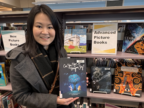 "A portrait of Viola Hernandez smiling in a dark coat and scarf while holding a copy of her book, 'Can You Hear the Halcyons Sing?' She is standing in front of library bookshelves displaying various children's books and signs for 'New Picture Books' and 'Advanced Picture Books'."